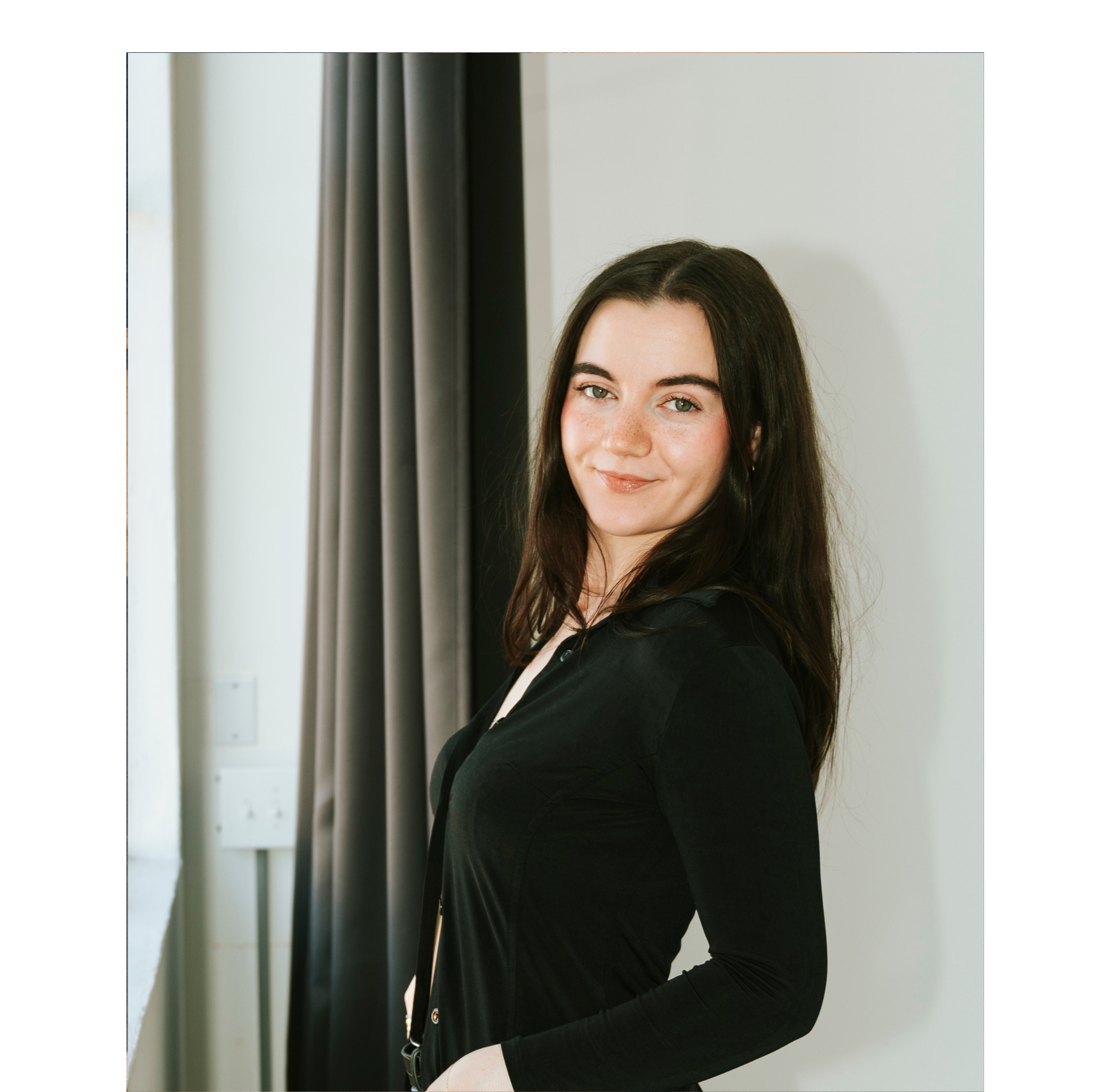 Woman in a black jacket standing against a white wall with curtains.