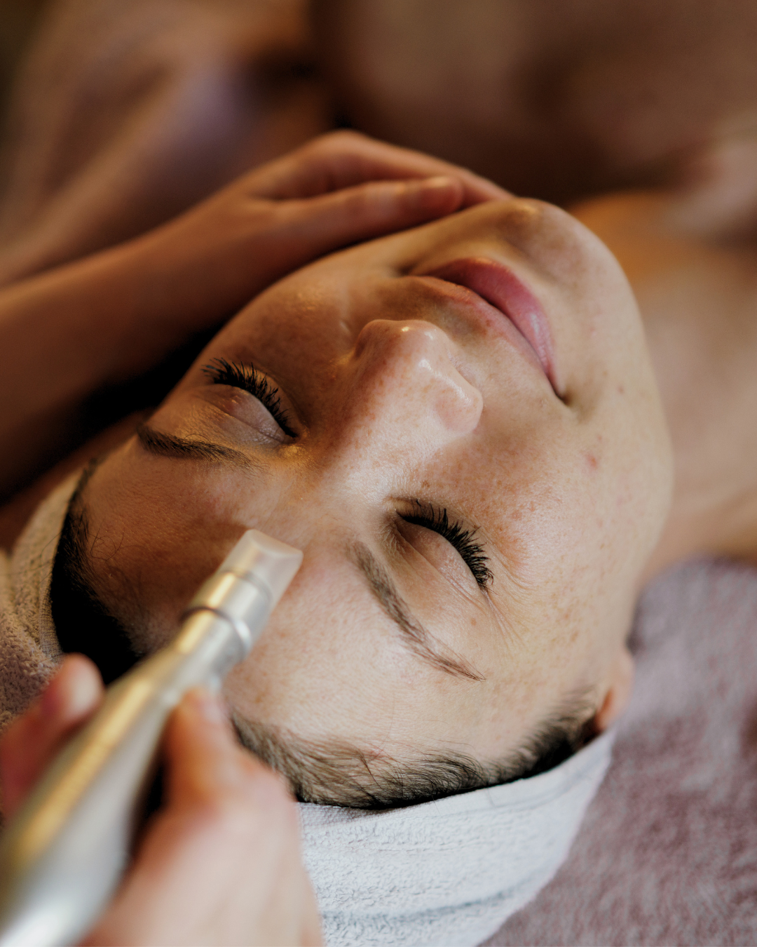 A close-up lifestyle shot of a woman receiving a facial treatment, with a therapist's hand holding a metallic device against her forehead. She has a white towel wrapped around her head, and the background is softly blurred.