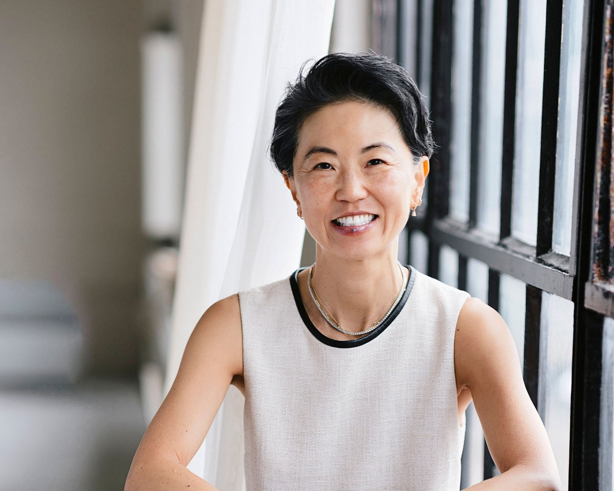 A smiling woman with short dark hair wearing a sleeveless light beige top with a black collar trim, standing indoors near a window with white curtains and dark window bars.