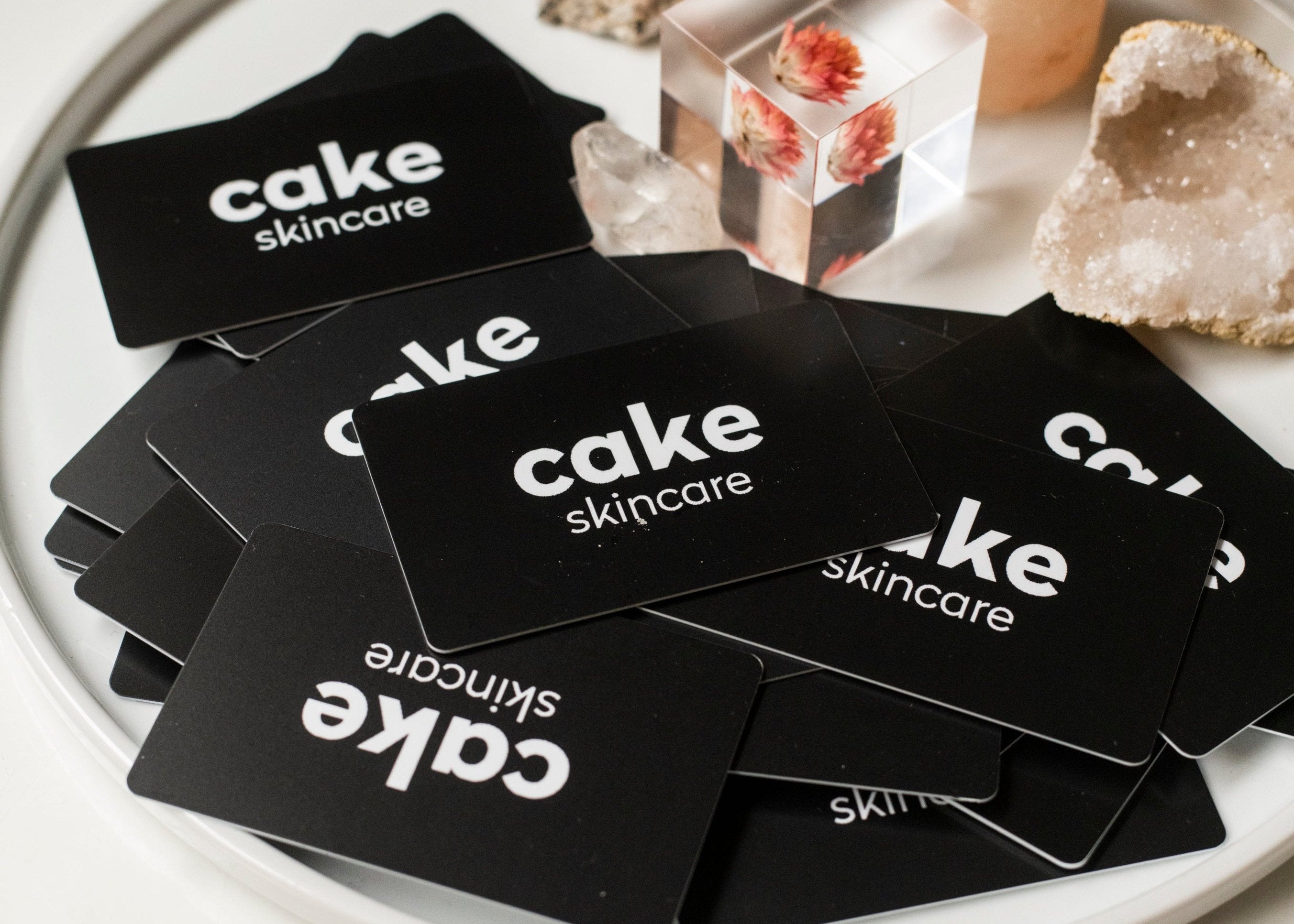 A stack of black gift cards with 'cake skincare' printed in white text, placed on a white plate with various crystals and a pink candle in the background.