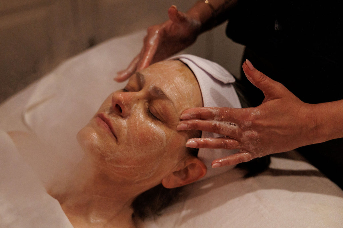 A close-up lifestyle shot of a woman receiving a facial treatment, with steam rising from a treatment bed while another person massages her face. She is wearing a white headband and draped in white towels.