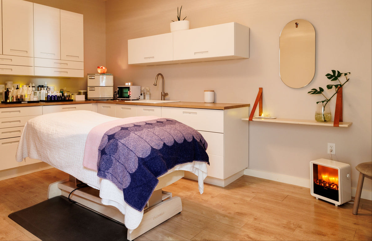 A modern spa treatment room featuring a white treatment table with a purple and blue scalloped blanket, white cabinetry, a built-in sink, a floating shelf with decorative items, and a small electric fireplace on the floor.
